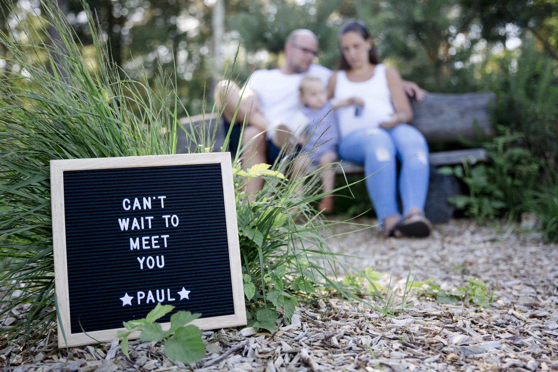 Babybauch-Shooting: Werdende Eltern und große Schwester sitzen auf Bank hinter Schild mit Aufschrift 'Can’t wait to meet you Paul