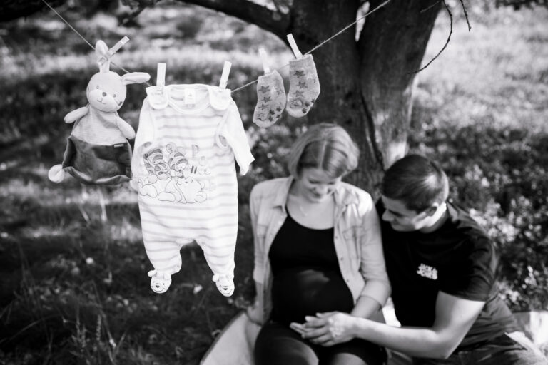 Schwarz-Weiß Babybauchfotografie: Werdende Eltern sitzen unter einem Baum, er streichelt den Bauch, Babykleidung im Vordergrund