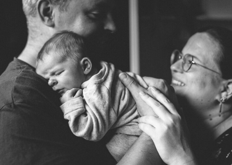 Schwarz-Weiß Newbornfoto: Baby schläft geborgen an Papas Schulter, Hände der Eltern halten das Kind